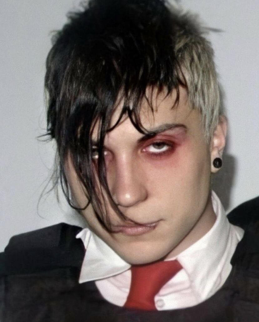 A picture of Frank Iero with bleached shaved sides of his hair and a black fringe hanging in front of his face reaching up to his lips. His eyes are droopy and he looks like he’s high/ mid-blink. He has red eyeshadow on and is wearing a dress shirt with a